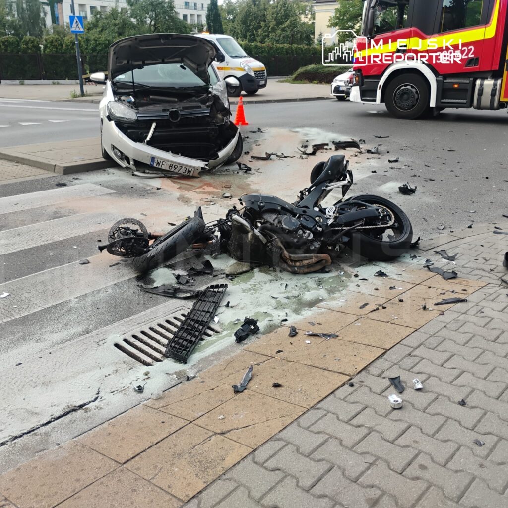 Zniszczony motocykl leży na jezdni obok rozbitego samochodu osobowego, wokół rozrzucone fragmenty pojazdów.