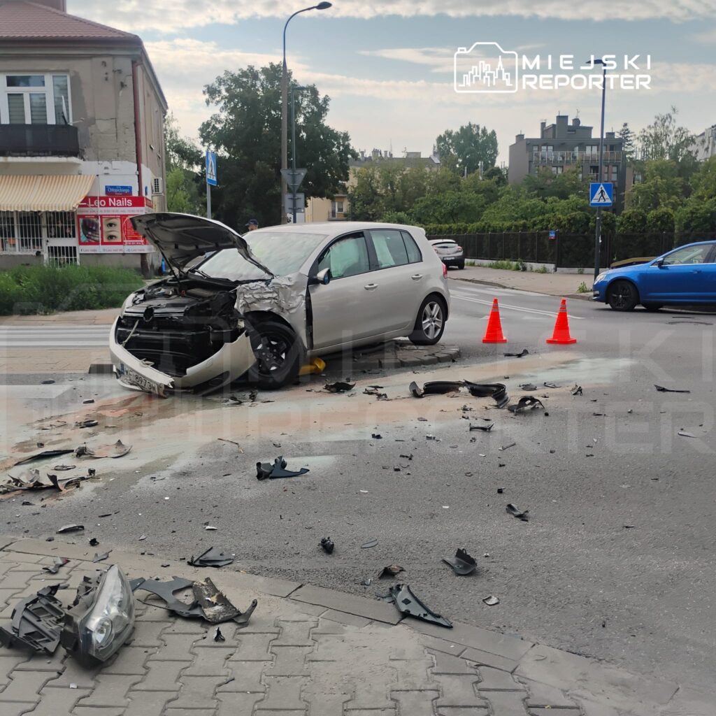 Rozbity samochód osobowy z otwartą maską leży na drodze, wokół rozsypane części pojazdu, a obok trzy pomarańczowe pachołki.