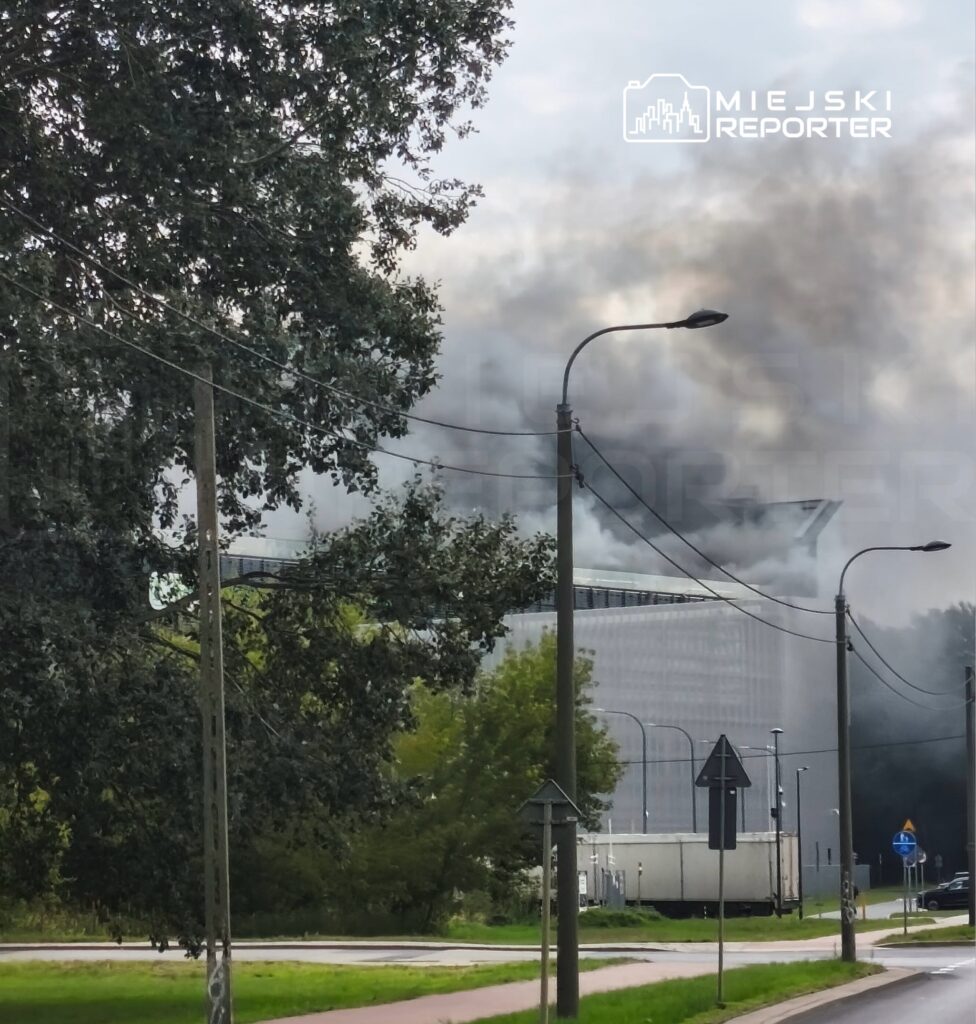 Gęsty dym wydobywający się z nowoczesnego budynku w miejskim otoczeniu, otoczony drzewami i latarniami ulicznymi.