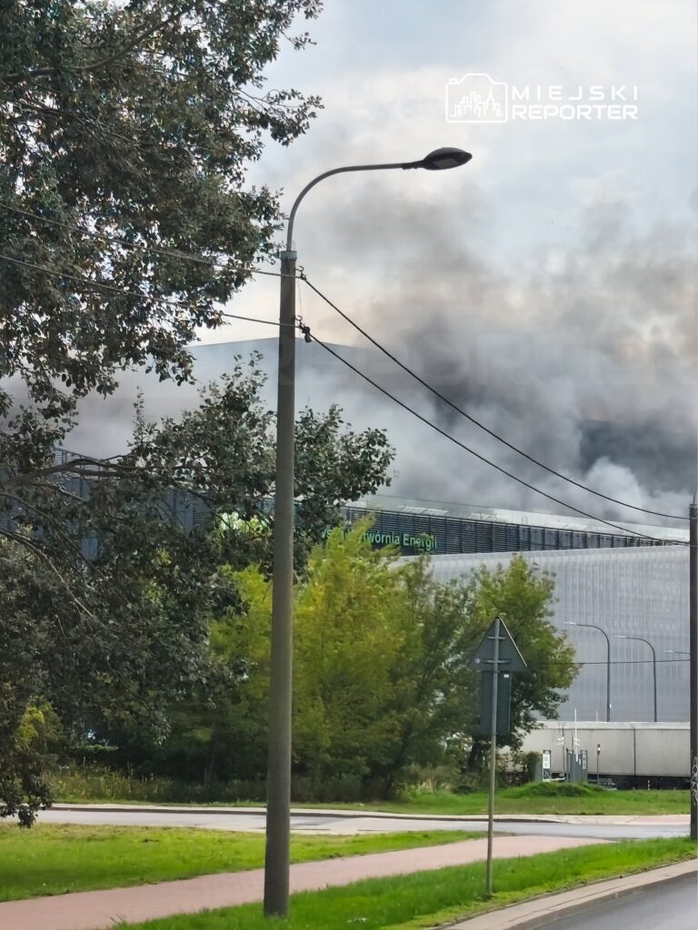 Gęsty dym unoszący się nad nowoczesnym budynkiem elektrowni, otoczonym drzewami i latarniami ulicznymi.