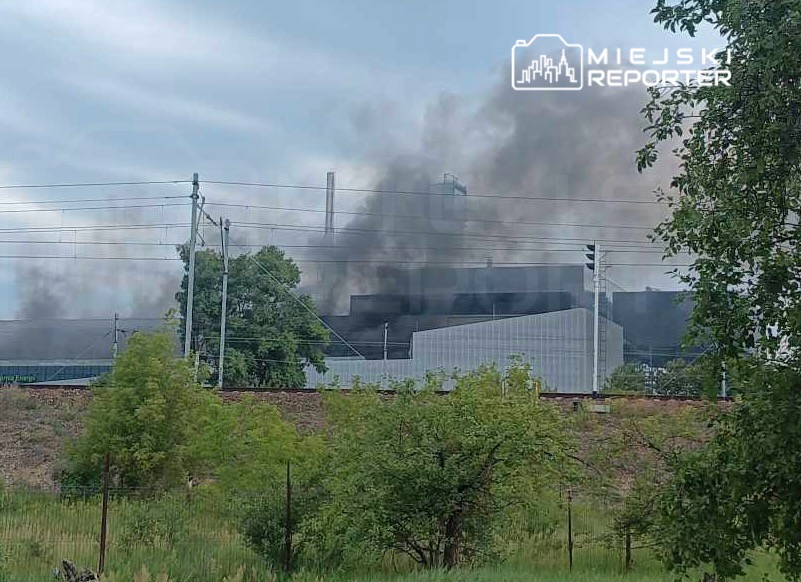 Z zakładu przemysłowego wydobywa się gęsty dym, otoczony zielonymi drzewami i liniami energetycznymi.