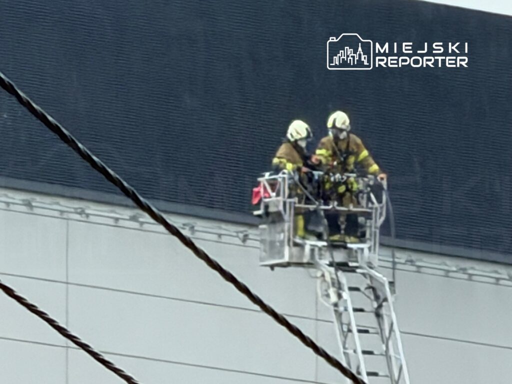 Dwaj strażacy w specjalnych hełmach pracują na podnośniku, wykonując zadania na elewacji budynku.
