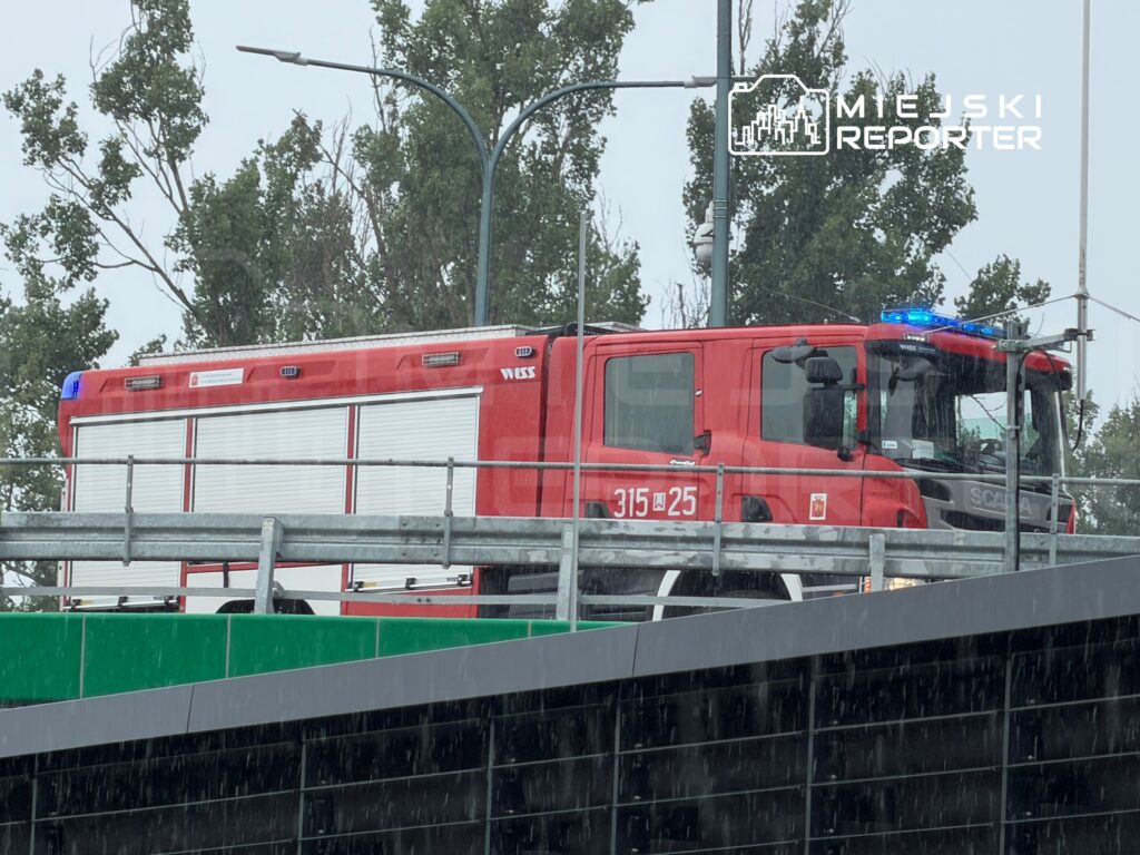 Czerwony wóz strażacki z niebieskimi światłami stoi na parkingu, w tle drzewa w deszczu.