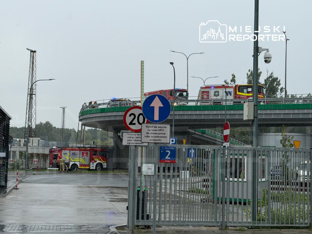 Strażacy przy wozie strażackim na parkingu pod wjazdem na rondo, w tle widoczny deszczowy krajobraz.