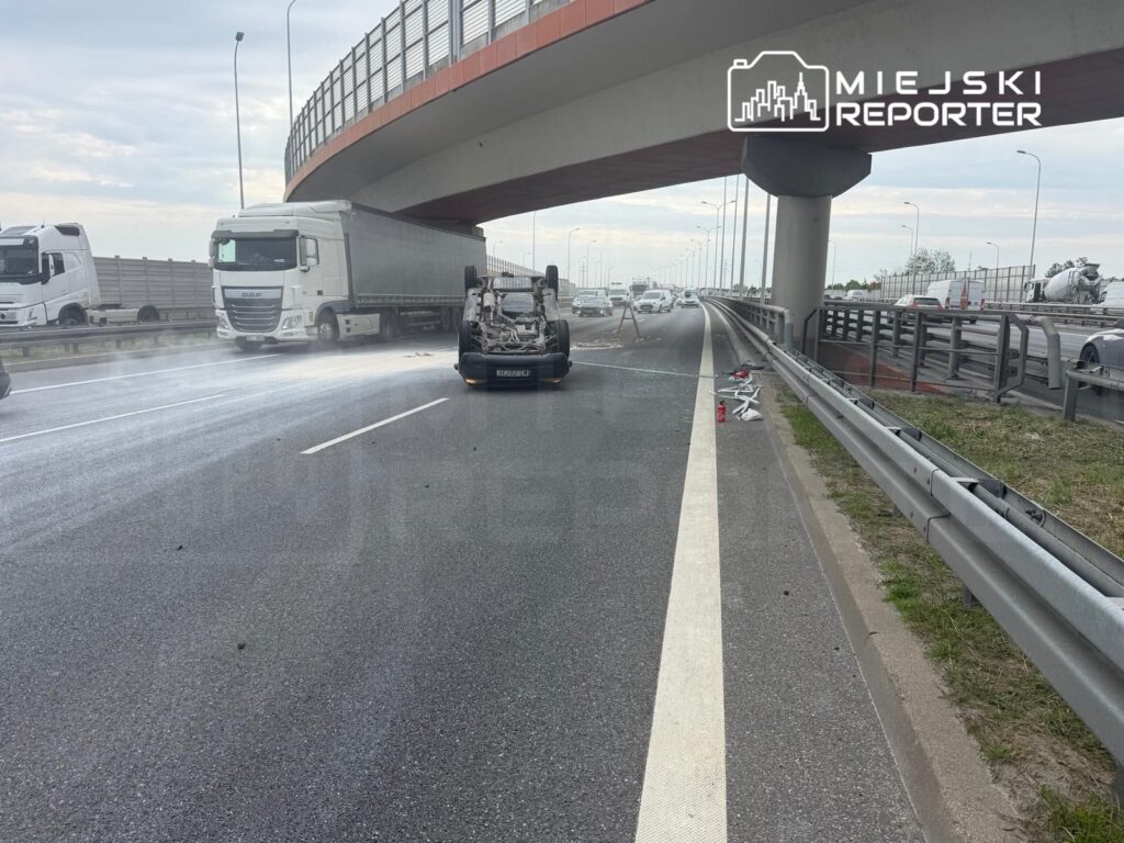 Na drodze leży przewrócony samochód osobowy, obok niego kilka ciężarówek. W tle widoczna jest estakada.