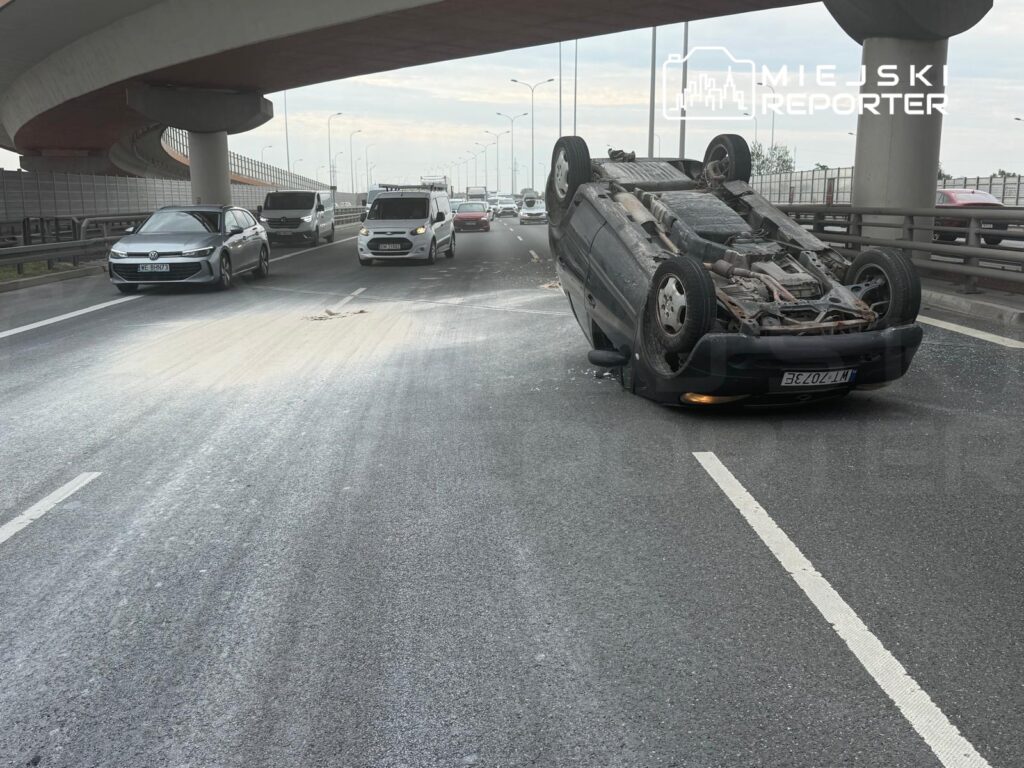 Na drodze leży przewrócone auto, obok poruszają się inne pojazdy, a na jezdni widoczne są ślady zdarzenia.