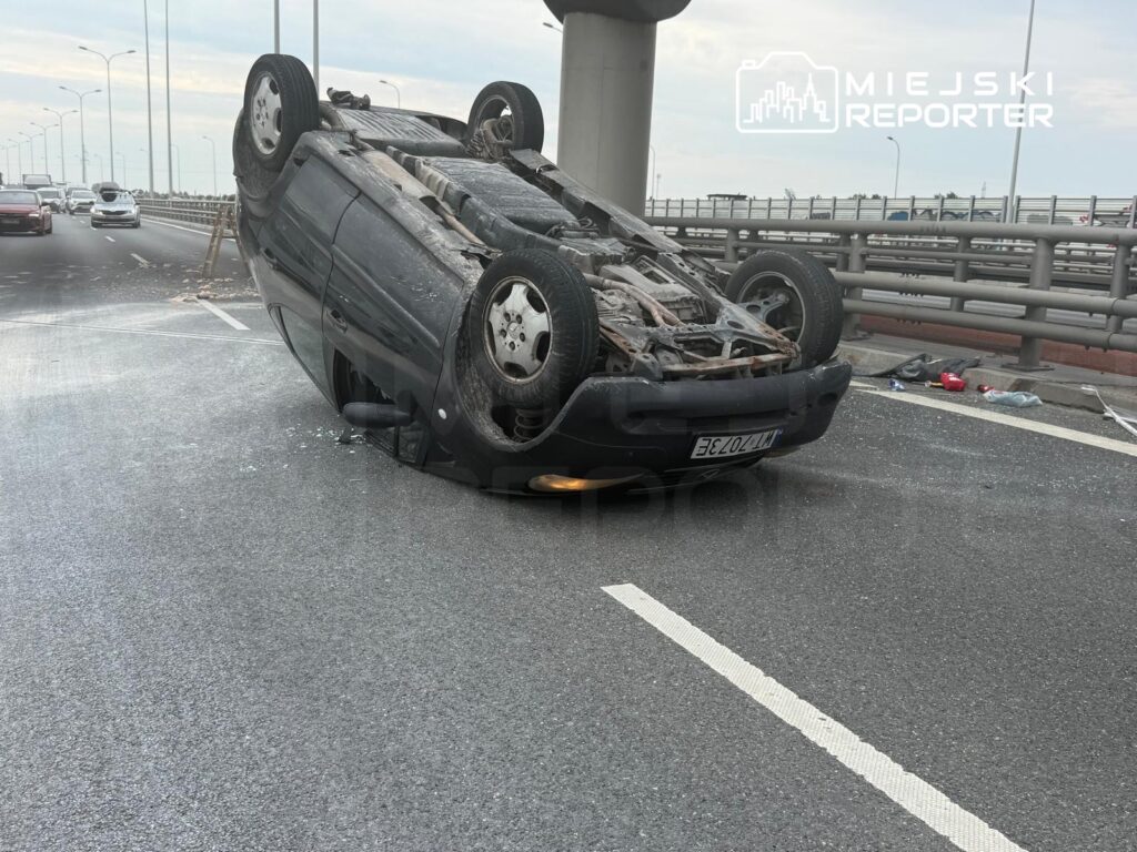 Czarny samochód osobowy leży na dachu na pasie drogowym, wokół widoczni inni kierowcy i fragmenty szkła.