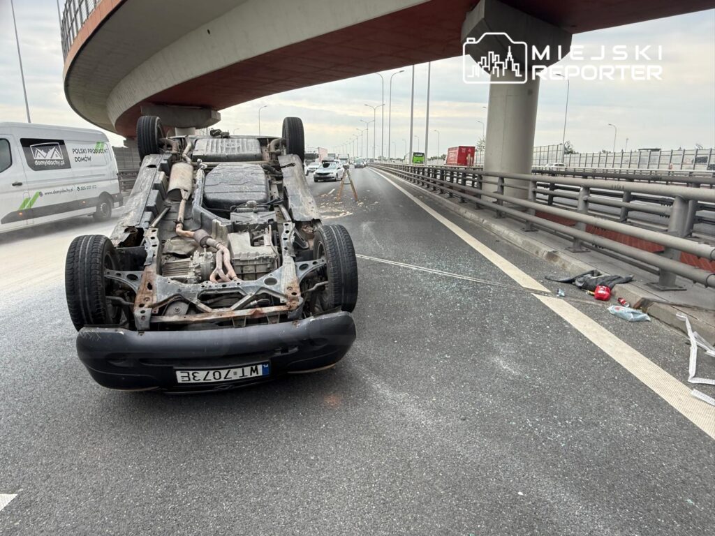 Przewrócone auto leży na jezdni pod wiaduktem, wokół rozbite szkło i pozostałości zderzenia.
