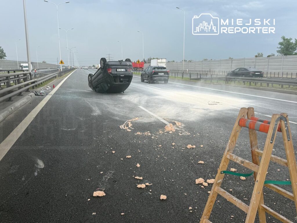 Na drodze leży przewrócone auto, obok widoczny jest sprzęt budowlany i rozrzucone resztki materiału.
