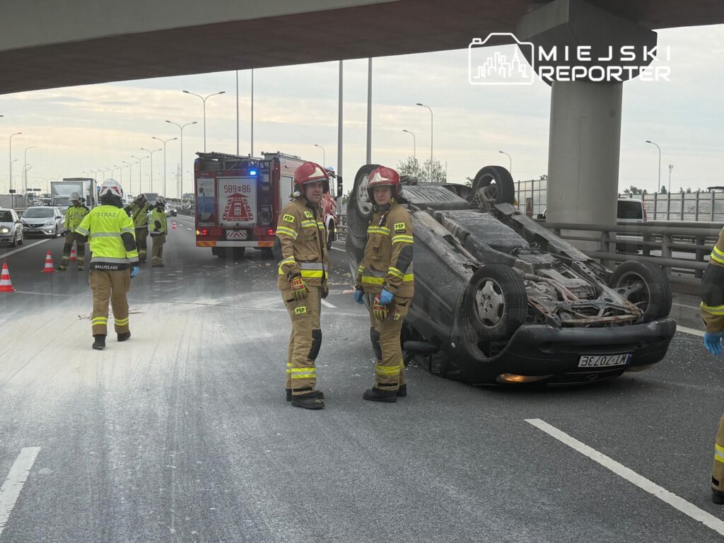 Strażacy w mundurach z logo PSP zabezpieczają miejsce wypadku na drodze, gdzie leży przewrócone auto.