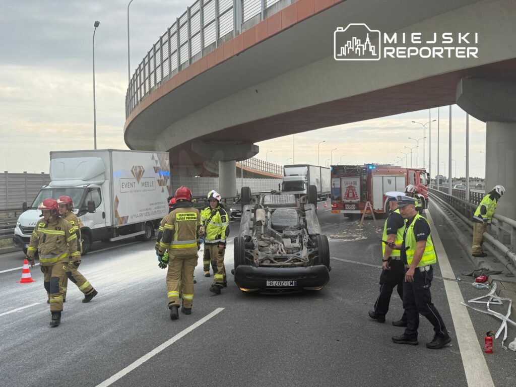 Grupa strażaków i policjantów pracuje przy przewróconym samochodzie na drodze, obok stoi ciężarówka.