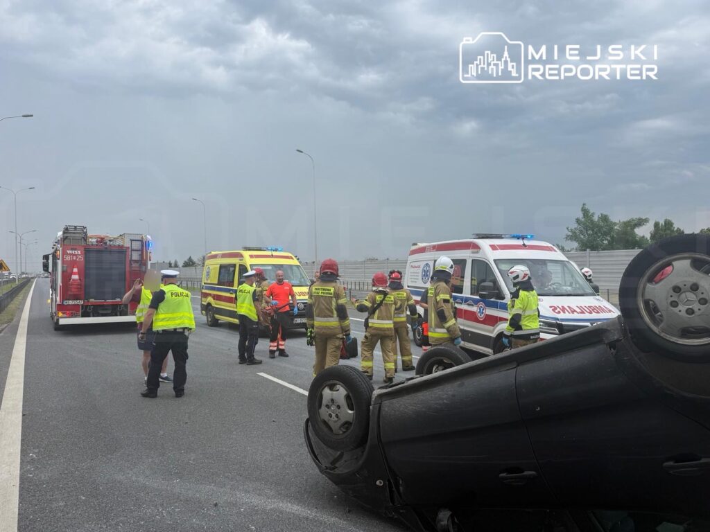 Grupa ratowników i policjantów otacza przewrócone na dach auto na drodze, obok stoją pojazdy straży pożarnej i karetka.
