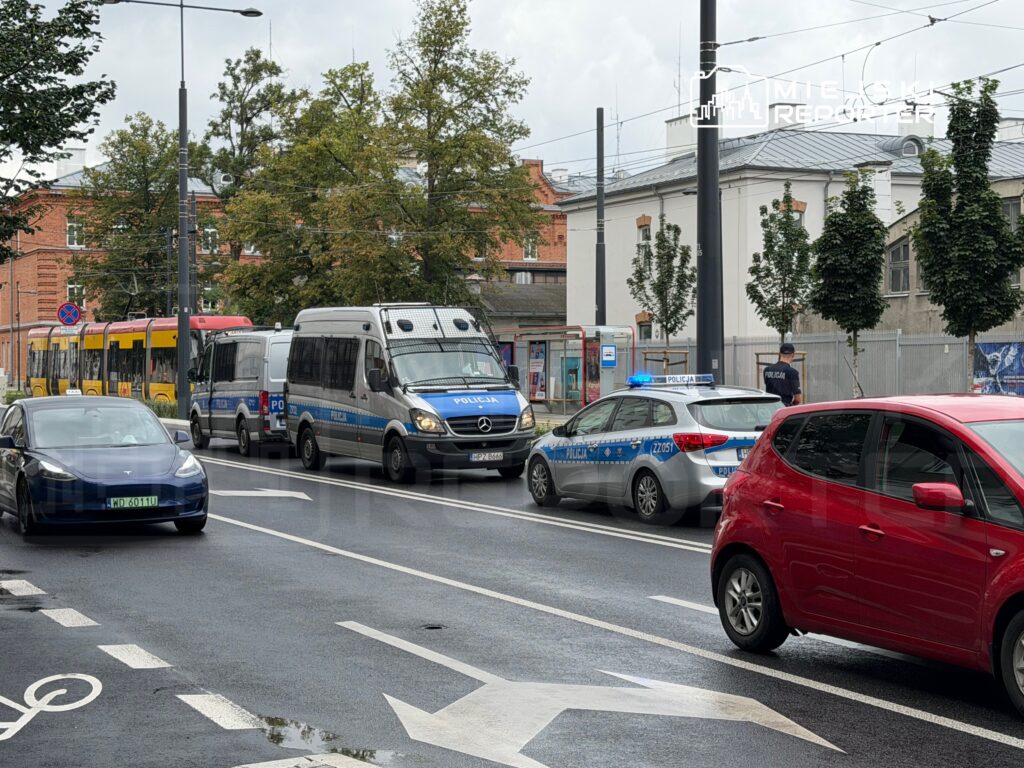 Policyjny radiowóz z włączonymi sygnałami świetlnymi zatrzymany na ruchliwej ulicy, obok stojącego tramwaju.