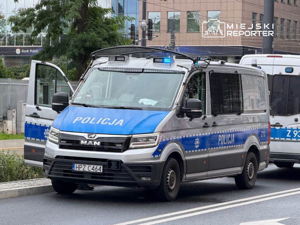 Radiowóz policyjny z niebieskimi światłami zaparkowany na ulicy obok nowoczesnego budynku banku.