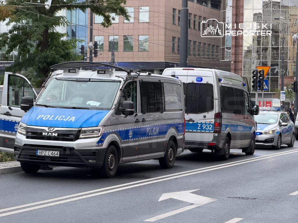 Policyjny pojazd man oraz furgonetka z oznaczeniem Z 932 zatrzymane na ulicy, w tle nowoczesne budynki.