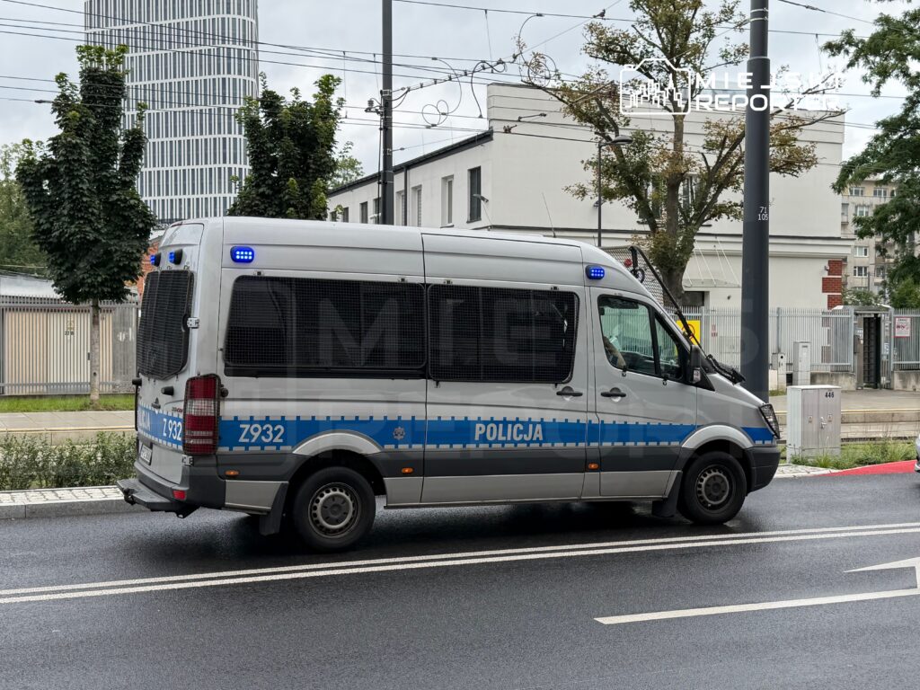 Policyjny radiowóz z niebieskimi światłami zaparkowany na ulicy obok nowoczesnego biurowca.