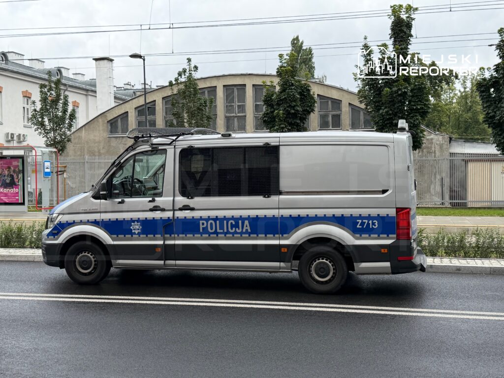 Policyjny furgon z niebieskimi oznaczeniami stoi na ulicy obok przystanku autobusowego.
