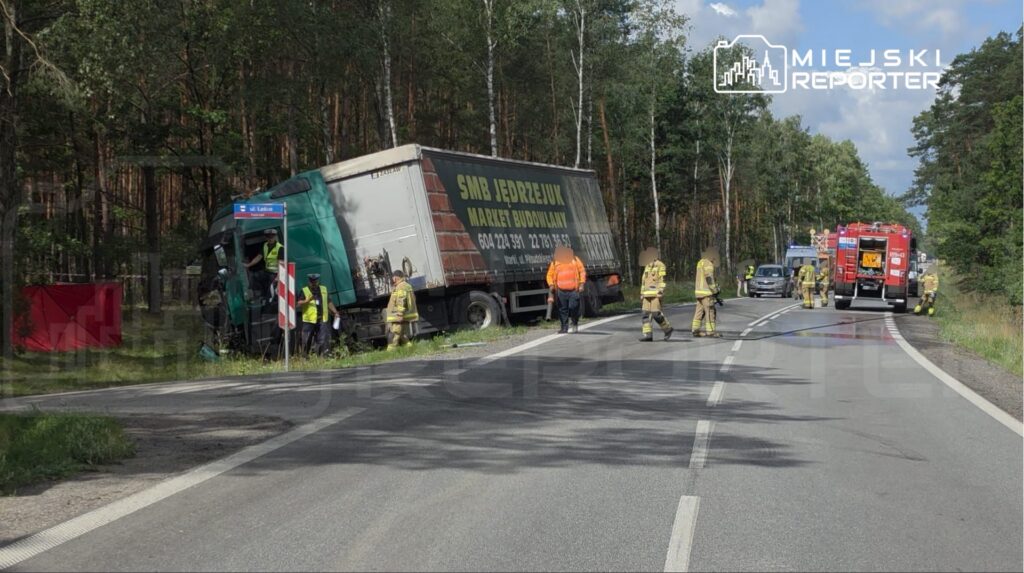 Wielu strażaków oraz służby ratunkowe pracują przy przewróconej ciężarówce na poboczu drogi, otoczonej lasem.