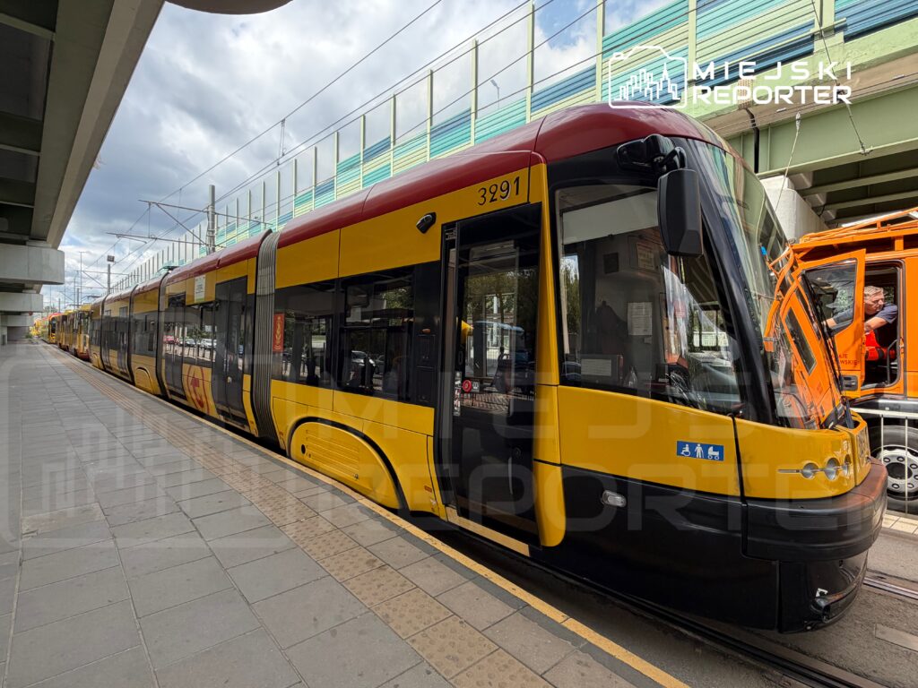 Tramwaj w żółto-czerwonym kolorze zatrzymany na przystanku, obok widać pracowników w pomarańczowych kamizelkach.