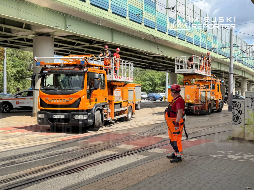 Pracownicy w pomarańczowych uniformach obsługują podnośnik koszowy przy torach tramwajowych, obok ruchliwej ulicy.