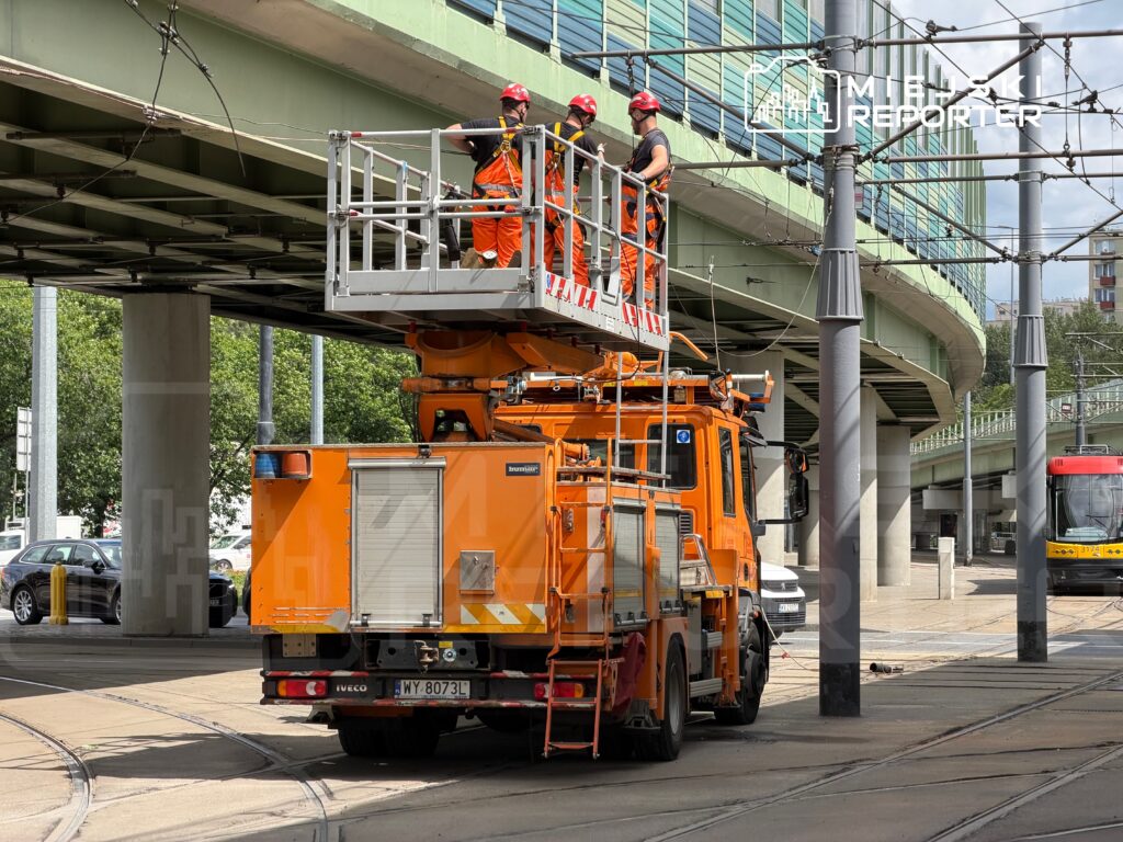 Pracownicy w pomarańczowych kombinezonach wykonują prace przy trakcji tramwajowej z podnośnika umieszczonego na ciężarówce.