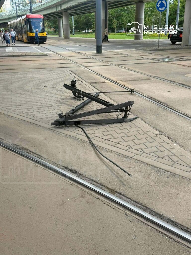 Uszkodzony element torów tramwajowych leży na chodniku obok przejazdu tramwajowego, w tle widoczny tramwaj.