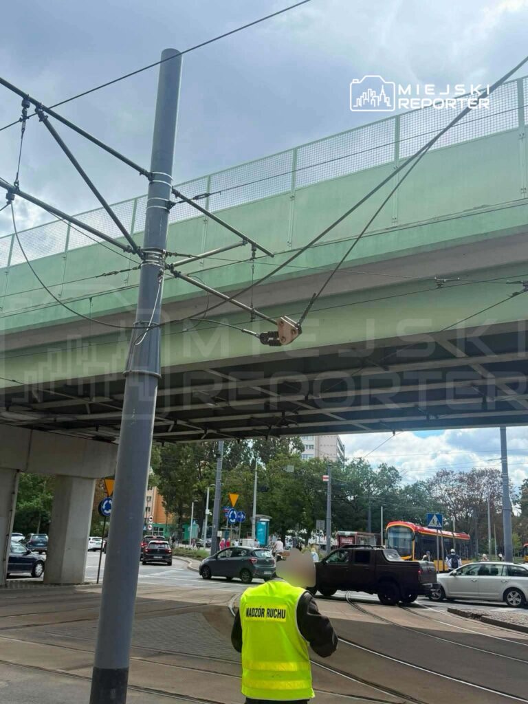 Mężczyzna w żółtej kamizelce nadzoru ruchu stoi na skrzyżowaniu, obserwując ruch tramwajowy i samochodowy.