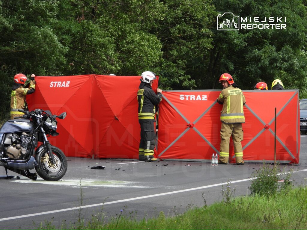 Czterech strażaków ustawia czerwony parawan na drodze, obok leży motocykl, a w tle widać zieleń.