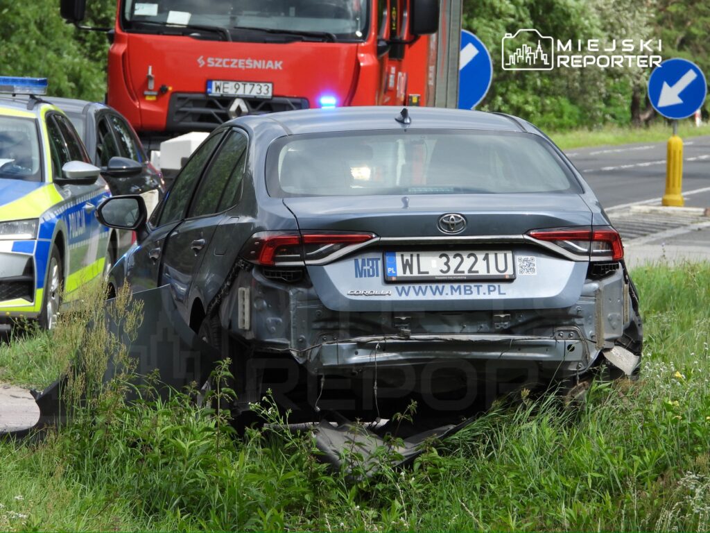 Uszkodzony samochód osobowy marki Toyota stoi na poboczu drogi, obok widać radiowóz policyjny i pojazd straży pożarnej.