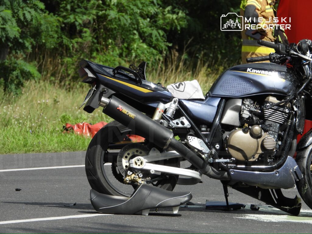 Motocykl marki Kawasaki uszkodzony na drodze, obok leży odcięta część wydechu, w tle zieleń i pas drogowy.