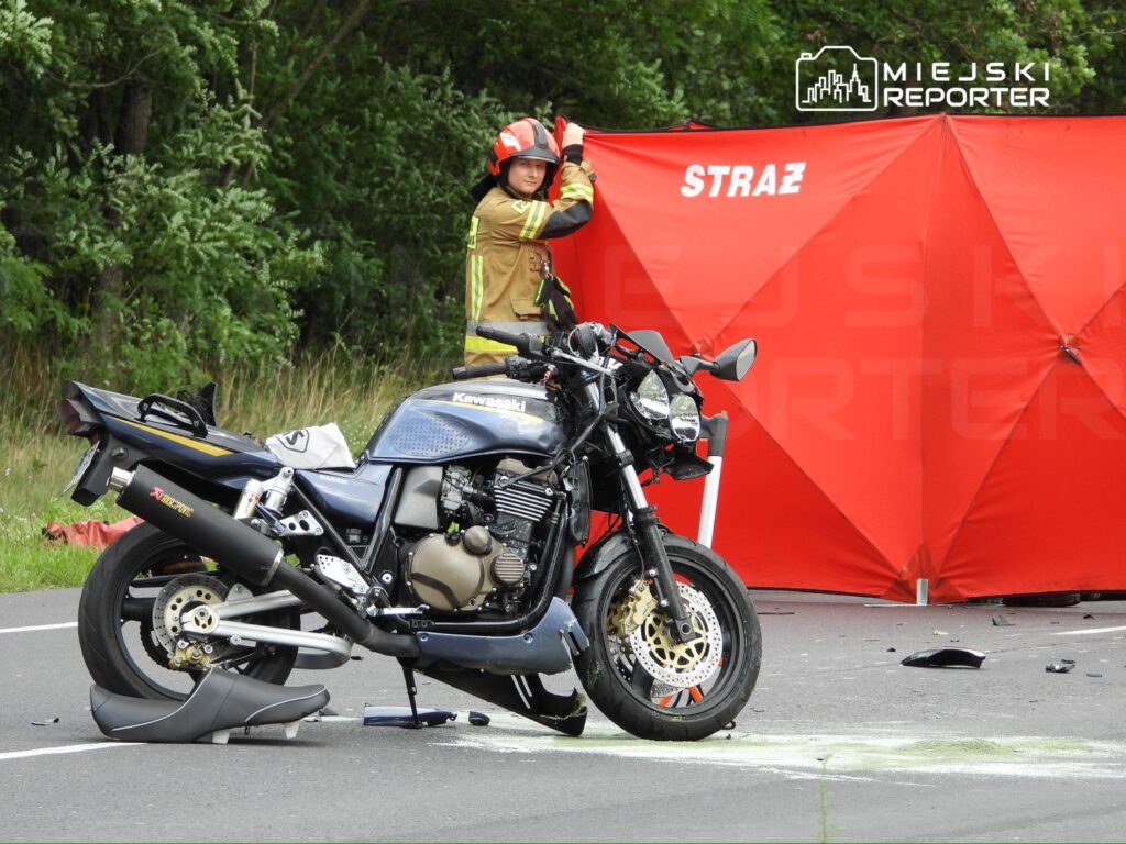 Strażak ustawiający czerwony namiot ochronny obok przewróconego motocykla na drodze, w otoczeniu zieleni.