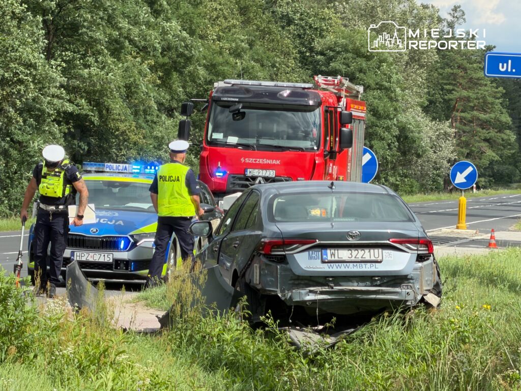 Policjanci w odblaskowych kamizelkach badają wypadek drogowy obok uszkodzonego samochodu na poboczu.