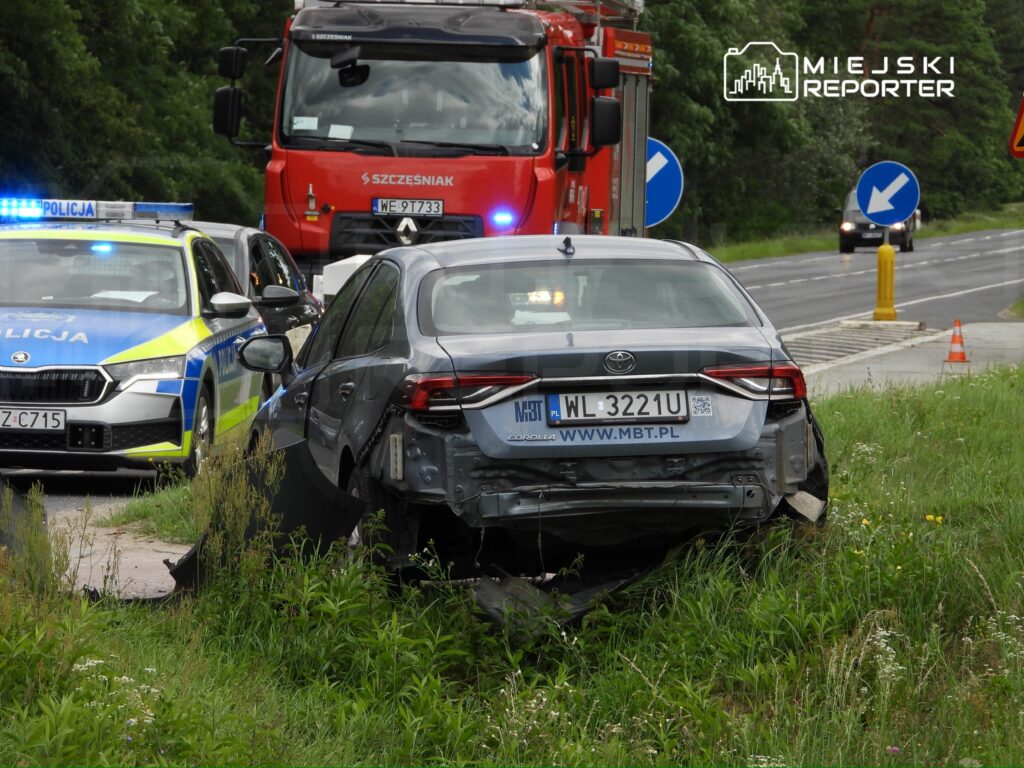 Uszkodzony samochód osobowy zaparkowany na trawie, obok policyjny radiowóz oraz wóz strażacki, w tle droga.