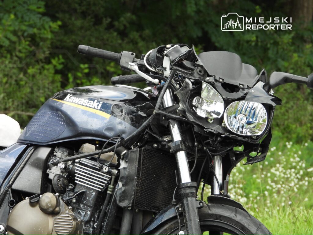 Zniszczony motocykl marki Kawasaki stoi na trawie, z uszkodzoną przednią częścią i odłamanymi reflektorami.