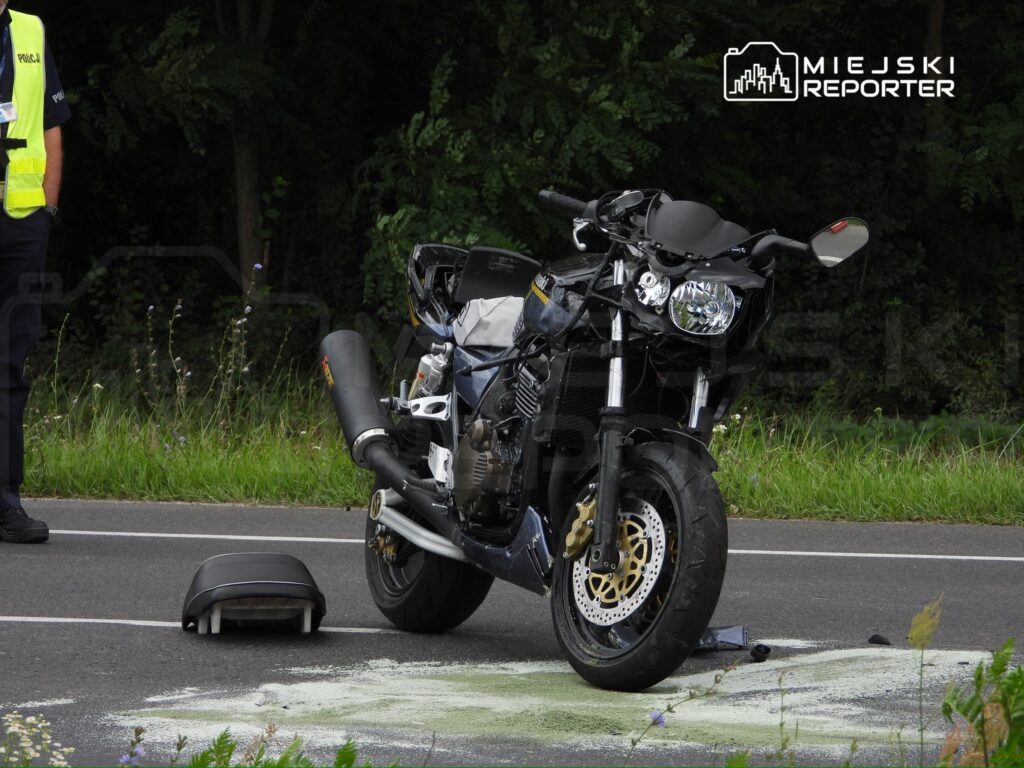 Motocykl leży na jezdni, obok stoi policjant w kamizelce odblaskowej, a na asfalcie widoczne ślady.