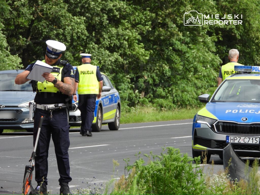 Policjant z notatnikiem bada miejsce zdarzenia na drodze, obok stoi radiowóz policyjny.