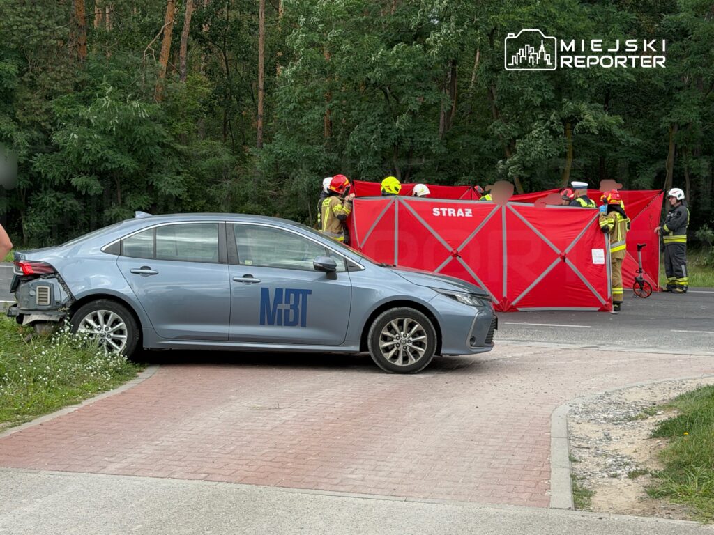 W miejscu wypadku drogowego strażacy pracują przy czerwonym namiocie, obok stoi uszkodzony samochód.