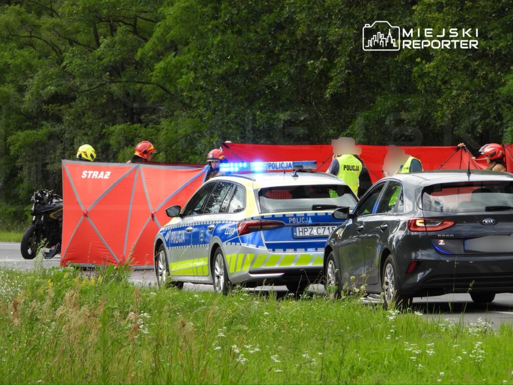 Na drodze zatrzymane pojazdy, z policyjnym radiowozem i motocyklowym, obok strażacy zabezpieczający miejsce zdarzenia.