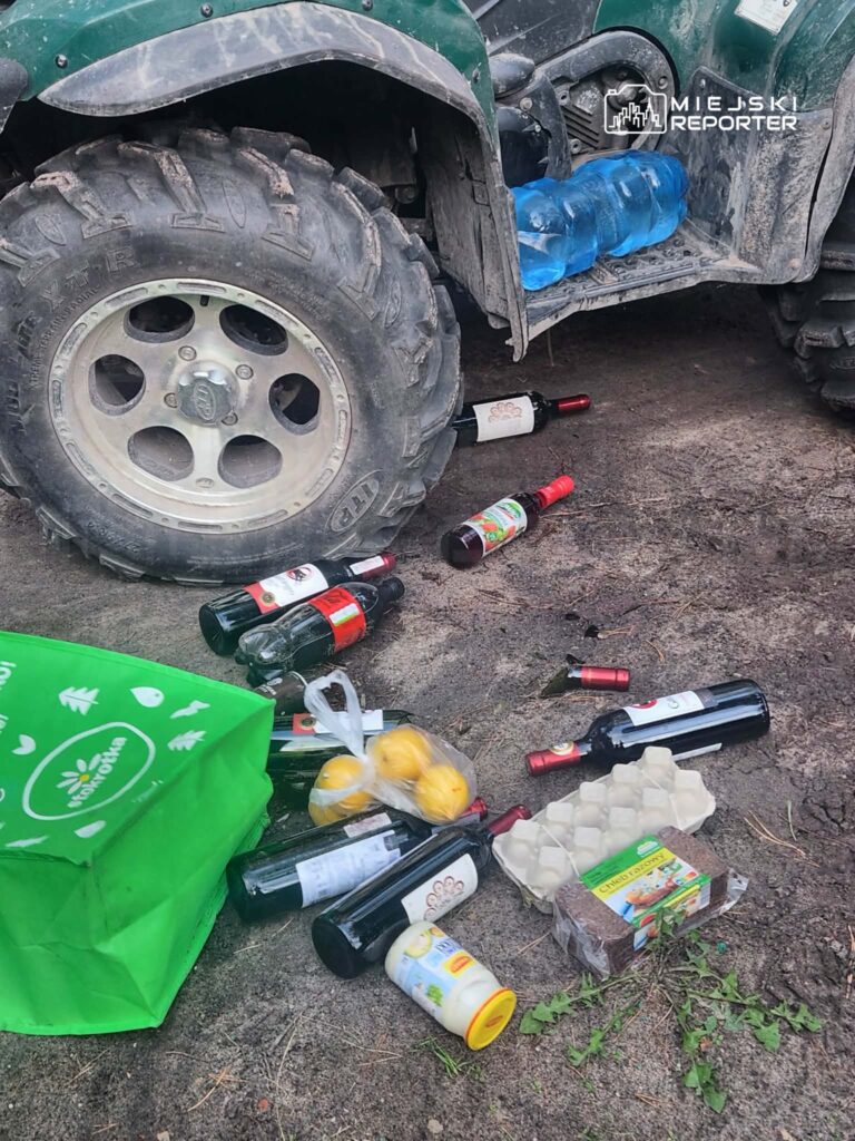 Na ziemi leżą porozrzucane butelki wina, cytryny, opakowania po jedzeniu oraz torba na zakupy obok koła pojazdu terenowego.