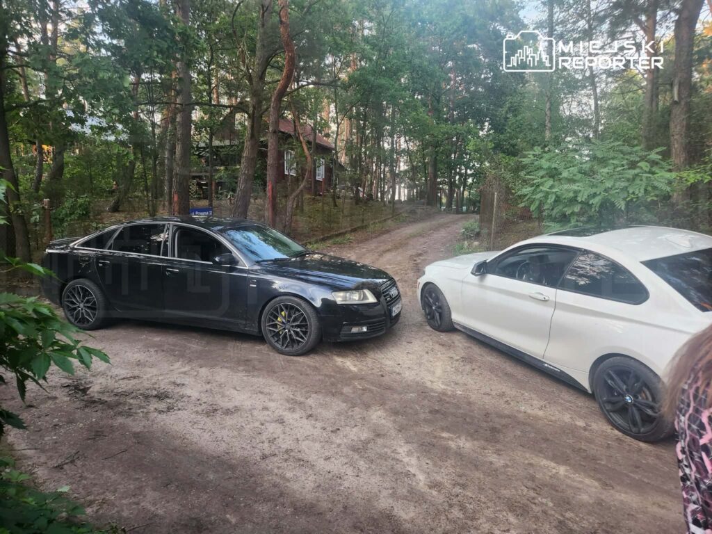 Czarne i białe samochody osobowe zaparkowane na leśnej drodze w otoczeniu drzew.