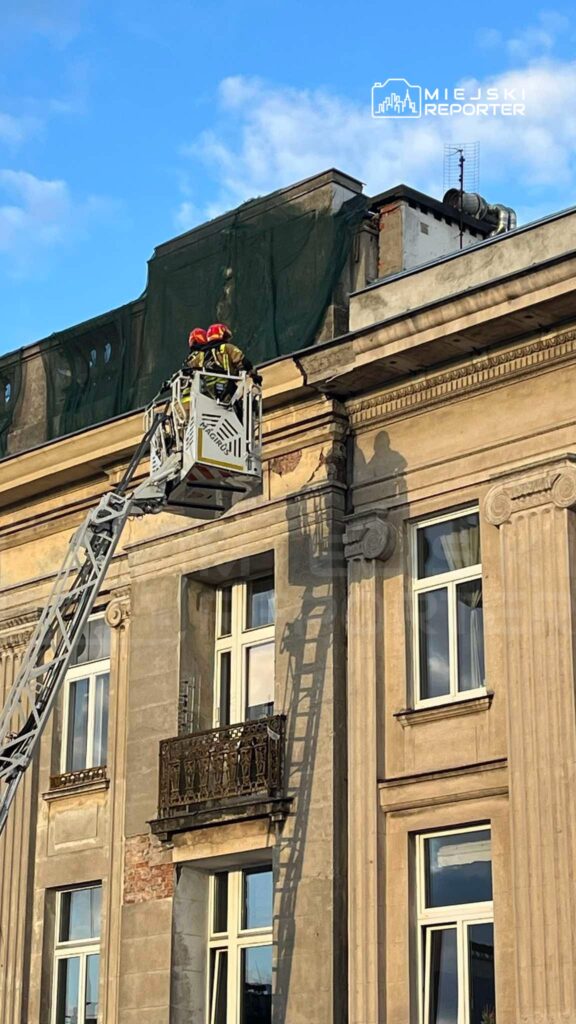 Dwaj strażacy w kaskach pracują na wysięgniku, naprawiając elewację budynku z przeszkloną fasadą.