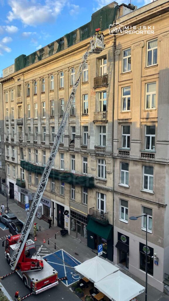 Ratownicy w hełmach pracują na dachu budynku, korzystając z podnośnika strażackiego w miejskiej scenerii.