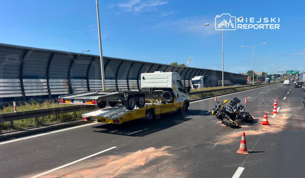 Wydarzenie drogowe z motocyklem leżącym na jezdni, obok stoi laweta z podnoszoną platformą. Na drodze ustawione pachołki ostrzegawcze.
