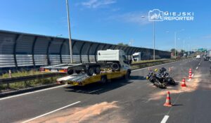 Wydarzenie drogowe z motocyklem leżącym na jezdni, obok stoi laweta z podnoszoną platformą. Na drodze ustawione pachołki ostrzegawcze.