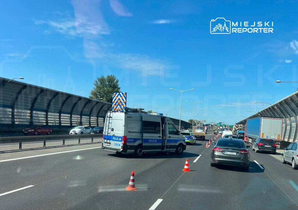 Karetka policyjna z włączonymi sygnałami blokuje pas ruchu na autostradzie, obok stoją samochody osobowe.
