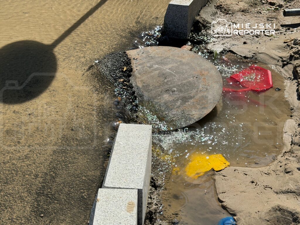 Woda spływa z odkrytej studzienki na mokrą ziemię, obok leżą czerwony i żółty plastikowy przedmiot.