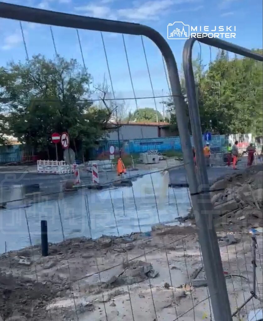 Robotnicy w odblaskowych kamizelkach pracują na zmodernizowanej drodze, otoczeni przez ogrodzenie budowlane.