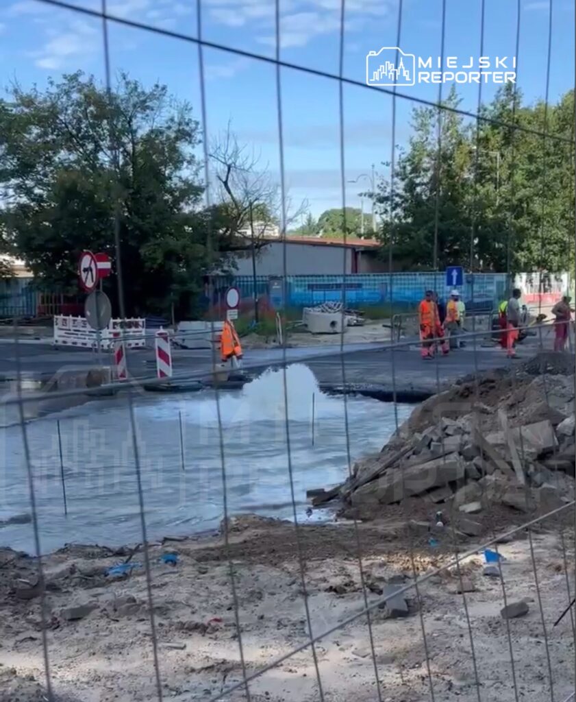Robotnicy w pomarańczowych kamizelkach pracują przy remoncie drogi, obok wytryskującej wody z uszkodzonego rurociągu.
