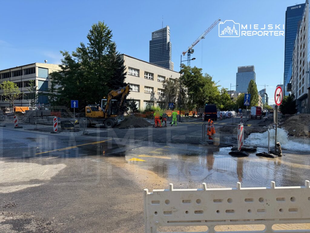 Robotnicy w pomarańczowych kamizelkach prowadzą prace budowlane na ulicy, obok koparki i wyciekającej wody.