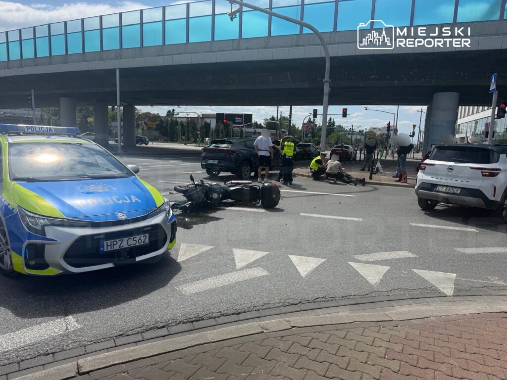 Na skrzyżowaniu leży przewrócony motocykl, a obok niego grupa osób oraz radiowóz policyjny.